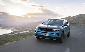 Türkis-schwarzer Opel Mokka Electric fährt bei Sonnenuntergang auf einer Küstenstraße, Berge und Meer im Hintergrund, Blick frontal von vorn.