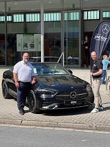 Zwei Männer stehen vor einem schwarzen Mercedes-Benz Coupé und lächeln in die Kamera. Rechts im Hintergrund ist eine Mercedes-Fahne, dahinter Glasfassade der Stadthalle.