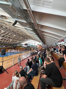 Innenaufnahme einer Sporthalle mit gefüllter Tribüne. Auf der Spielfläche stehen in Formation mehrere Sportlerinnen in weißen Anzügen bei einer Vorführung.