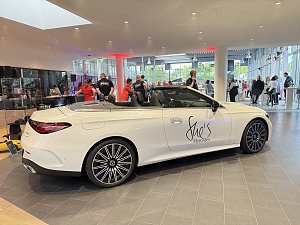 Ein wei&szlig;es Mercedes-Cabriolet mit der Aufschrift She�s Mercedes steht in einem Autohaus, im Hintergrund sind G&auml;ste und Personal in roten T-Shirts zu sehen.