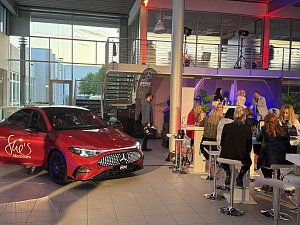 Roter Mercedes-Benz CLA 250+ steht in einer Veranstaltungshalle, im Hintergrund sitzen G&auml;ste an Stehtischen. Mercedes-Benz CLA 250+ mit EQ-Technologie | Energieverbrauch kombiniert: 14,1-12,2 kWh/100 km | CO2Emissionen kombiniert: 0 g/km | CO2-Emissionsklasse: A