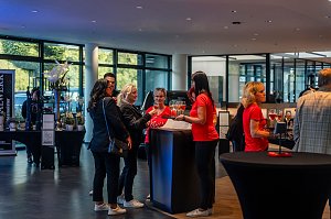 Mehrere Frauen, teils in roten She�s Mercedes-T-Shirts, teils in schwarzen Outfits, stehen an einem Stehtisch mit Gl&auml;sern und unterhalten sich in einem hellen Autohaus.