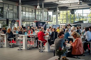 Publikum im Showroom: Viele Frauen sitzen an hohen wei&szlig;en Tischen in einem gro&szlig;en, hellen Autohaus mit Glasfront, trinken und unterhalten sich. Im Hintergrund sind Mercedes-Werbematerialien und Fahrzeuge sichtbar.