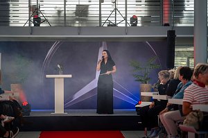 Christina Dost spricht auf einer B&uuml;hne mit Mercedes-Logo im Hintergrund, rotem Teppich davor und Publikum auf beiden Seiten.