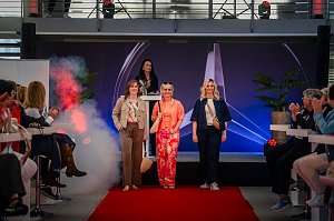 Drei Frauen laufen in modischen Outfits &uuml;ber einen roten Teppich auf einer B&uuml;hne mit Mercedes-Logo, w&auml;hrend eine Moderatorin am Pult steht. Das Publikum applaudiert.
