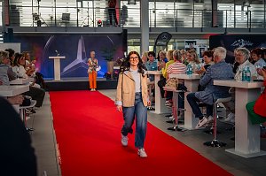 Eine junge Frau mit dunklen, langen Haaren, Brille, beiger Jacke, hellblauem Oberteil, blauen Jeans und wei&szlig;en Sneakern l&auml;uft bei einer Modenschau &uuml;ber einen roten Teppich.