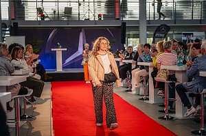 Eine Frau mit lockigem, blondem Haar, beiger Jacke, wei&szlig;em Oberteil, Leopardenmuster-Hose und kleiner schwarzer Umh&auml;ngetasche l&auml;uft &uuml;ber den roten Teppich.