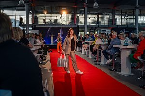Frau im beigefarbenen Hosenanzug mit braunem Oberteil und gro&szlig;em, silberfarbenem Shopper l&auml;uft auf dem roten Teppich.