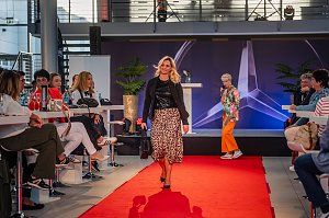  Frau mit blonden Haaren, schwarzem Oberteil, schwarzer Jacke und Leopardenrock tr&auml;gt eine schwarze Handtasche &uuml;ber den Laufsteg.