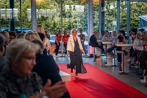 Frau mit kurzem blondem Haar, wei&szlig;er Jacke und langem, dunkelblauem Kleid mit wei&szlig;en Punkten l&auml;uft &uuml;ber den roten Teppich.
