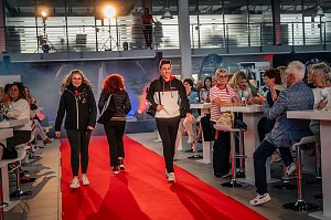 Mehrere Models in sportlicher Kleidung � schwarze Jacken mit roten und wei&szlig;en Akzenten � laufen gleichzeitig &uuml;ber den roten Teppich, ein junger Mann und eine junge Frau vorne, weitere Personen dahinter.