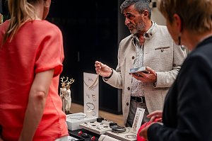 Ein Mann in heller Jacke pr&auml;sentiert Schmuckst&uuml;cke an einem Stand, w&auml;hrend zwei Frauen interessiert zuh&ouml;ren. Auf dem Tisch liegen Ketten, Armb&auml;nder und Ringe.
