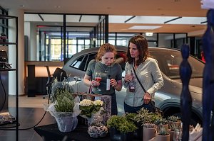 Zwei Frauen betrachten und riechen Pflanzen an einem Floristik-Stand im Autohaus, eine h&auml;lt ein Glas mit rosafarbenem Getr&auml;nk.