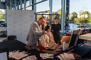 Friseurmeister in grauem Anzug h&auml;lt die Haare einer sitzenden Frau und schl&auml;gt ihr eine Frisur vor, w&auml;hrend zwei weitere Frauen zusehen. Vor ihnen stehen Kosmetik- und Styling-Utensilien auf Tischen.