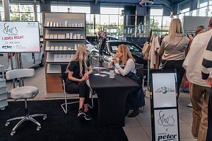 Zwei Frauen sitzen an einem Tisch und unterhalten sich; im Hintergrund steht ein Regal mit Kosmetikprodukten und ein Bildschirm mit der Aufschrift She�s Mercedes Ladies Night 22. Mai 2025. Daneben st&ouml;bern Besucher an einem Kleiderst&auml;nder.