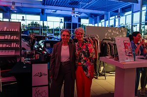 Zwei Frauen stehen im Autohaus vor einem Kleiderst&auml;nder und l&auml;cheln in die Kamera, links eine in grauem Hosenanzug, rechts Mandy Kossira in bunter Jacke mit Blumenmuster.
