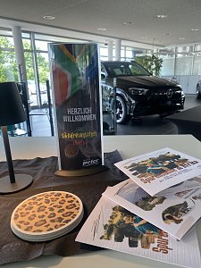 Dekoration zum Südafrikanischen Abend im Autohaus Peter: Ein Aufsteller mit der Aufschrift �Herzlich Willkommen zum Südafrikanischen Abend� und südafrikanischer Flagge, daneben Flyer mit Safari-Motiven und ein Untersetzer mit Leopardenmuster. Im Hintergrund steht ein schwarzer Mercedes im Showroom.