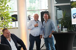 Afrikanischer Abend im Autohaus Peter: Helmut Peter und Abenteuer-Guide Hannu begrüßen die Gäste lachend, während Helmut Peter das Mikrofon hält. Links im Bild lacht ein weiterer Teilnehmer, rechts ist ein Stehtisch mit südafrikanischer Dekoration und ein Bildschirm mit einem Safari-Motiv zu sehen.