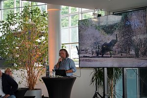 Abenteuer-Guide Hannu beim afrikanischen Abend im Autohaus Peter in Heiligenstadt: Er berichtet am Rednerpult von seinen Safari-Erlebnissen, während auf einem großen Bildschirm ein Elefant in der afrikanischen Savanne zu sehen ist.