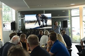 Abenteuer-Guide Hannu präsentiert beim südafrikanischen Abend im Autohaus Peter in Heiligenstadt spannende Eindrücke einer Safari, während auf dem Bildschirm ein Elefant zu sehen ist und die Gäste aufmerksam zuhören.