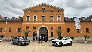 Vorderansicht des historischen Geb&auml;udes mit orangefarbener Fassade. Zwei Mercedes-Benz Fahrzeuge stehen auf dem Platz davor, w&auml;hrend sich einige Besucher am Eingang unterhalten. Rechts ist ein Banner mit Werbung f&uuml;r ein kulturelles Event zu sehen.