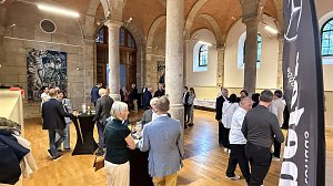 Innenaufnahme eines historischen Saals mit hohen Steins&auml;ulen und Holzboden. G&auml;ste stehen an Stehtischen, unterhalten sich und genie&szlig;en Getr&auml;nke. Im Hintergrund ist ein Buffet aufgebaut.