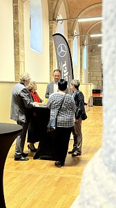 Eine kleine Gruppe von f&uuml;nf Personen unterh&auml;lt sich im historischen Saal an einem schwarzen Stehtisch. Im Hintergrund sind hohe Steins&auml;ulen, W&auml;nde aus Naturstein und ein gro&szlig;es Mercedes-Benz Werbef&auml;hnchen zu sehen.