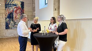 Vier Personen stehen und unterhalten sich an einem schwarzen Stehtisch in einem historischen Saal mit Holzboden und Steinw&auml;nden. Auf dem Tisch stehen Gl&auml;ser mit Sekt und Orangensaft. Im Hintergrund h&auml;ngt ein farbenfrohes Gem&auml;lde an der Wand.