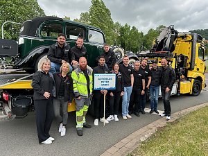 Gruppenfoto eines Teams des Autohaus Peter beim Festumzug in Sondershausen. Die Mitarbeitenden stehen vor einem klassischen Abschleppwagen des ADAC mit einem Oldtimer auf der Ladefläche. Alle tragen schwarze Autohaus-Peter-Bekleidung, einige lachen in die Kamera. Im Vordergrund wird ein Schild mit der Aufschrift Autohaus Peter GmbH gehalten. Im Hintergrund Bäume und bewölkter Himmel.