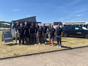 Gruppenfoto des Teams vom Autohaus Peter bei sonnigem Wetter. Zehn Personen stehen in schwarzer Teamkleidung mit Logo �Peter� nebeneinander vor einem silbernen Van und einem schwarzen Mercedes. Im Hintergrund sind ein Zelt, Werbebanner und weitere Fahrzeuge zu sehen. Alle lächeln in die Kamera.
