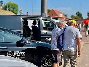 Zwei ältere Männer gehen nebeneinander über das Veranstaltungsgelände des Leichtathletik-Meetings Anhalt 2025, vorbei an schwarzen Mercedes-Benz Fahrzeugen vom Autohaus Peter, darunter ein Van mit geöffneter Schiebetür. Im Hintergrund sind weitere Besucher, Gebäude und ein sonniger, klarer Himmel zu sehen.