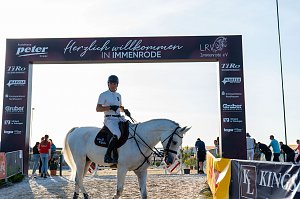 Ein Reiter in weißem T-Shirt und schwarzem Helm sitzt auf einem weißen Pferd unter einem Begrüßungsbogensorenn mit der Aufschrift �Herzlich willkommen in Immenrode�. Der Bogen ist mit Logos verschiedener Spo versehen. Im Hintergrund sind mehrere Personen und ein sonniger Himmel zu sehen.
