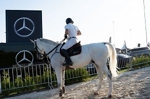 Ein Reiter in weißer Turnierkleidung sitzt auf einem weißen Pferd vor einem Mercedes-Benz Werbebanner mit zwei großen Logos. Der Reitplatz ist von Absperrgittern und grünen Sträuchern umgeben, im Hintergrund stehen Fahrzeuge und Eventzelte.