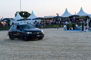 Ein schwarzer Mercedes-Benz SUV f&auml;hrt &uuml;ber den Sandplatz einer Reitanlage. Im Hintergrund sitzen zahlreiche Zuschauer unter wei&szlig;en Pagodenzelten und schwarzen Sonnenschirmen und verfolgen das Abendprogramm des Turniers. [Mercedes-AMG GLA 35 4MATIC | Energieverbrauch kombiniert: 9,3-8,8 l/100 km | CO2-Emissionen kombiniert: 211-200 g/km | CO2-Klasse: G]