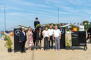 Siegerehrung beim Reitturnier in Immenrode: Ein Reiter sitzt auf seinem Pferd mit Siegerdecke und Blumenstrauß, daneben stehen fünf Erwachsene zur Gratulation. Die Szene spielt sich zwischen Mercedes-Benz-Hindernissen und unter wehenden Fahnen auf dem Turnierplatz ab.