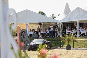 Zuschauer sitzen unter weißen Eventzelten an Tischen und verfolgen ein Springreitturnier. Vor den Zelten steht ein schwarzes Mercedes-Benz Cabrio, dahinter blühende Pflanzen und ein sonniger Himmel.
