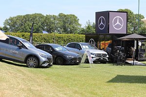 Drei Mercedes-Benz Fahrzeuge stehen auf einer grünen Wiese neben einem schwarzen Mercedes-Benz Cube. Vor dem Container befinden sich ein Sonnenschirm und ein kleiner Tisch mit Stühlen. Im Hintergrund sind Bäume und blauer Himmel zu sehen.