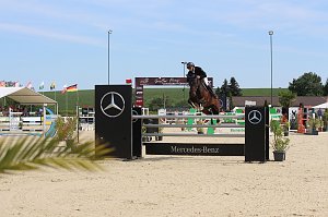 Ein Reiter in schwarzer Reitkleidung und Helm springt mit einem braunen Pferd über ein Hindernis mit Mercedes-Benz-Logo. Im Hintergrund sind Zuschauer unter einem weißen Zelt, mehrere Fahnen und eine grüne Landschaft zu sehen.