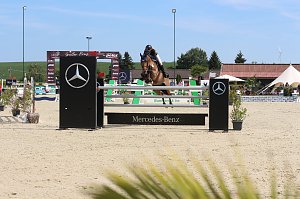 Eine Reiterin springt mit einem braunen Pferd über ein Mercedes-Benz-Hindernis auf dem Sandplatz beim Reitturnier in Immenrode. Im Hintergrund sind weitere Hindernisse, grüne Landschaft und weiße Zelte zu sehen.