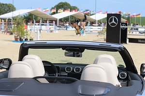 Blick über die Sitze eines schwarzen Mercedes-Benz Cabrios auf den Springplatz beim Reitturnier in Immenrode. Im Hintergrund springt ein Reiter über ein Hindernis, flankiert von Mercedes-Benz-Werbebannern und zahlreichen Länderflaggen.