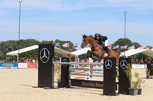 Ein Reiter springt mit einem braunen Pferd über ein Mercedes-Benz-Hindernis auf dem Sandplatz beim Reitturnier in Immenrode. Im Hintergrund sitzen Zuschauer unter weißen Tribünendächern, darüber wehen zahlreiche Länderflaggen.