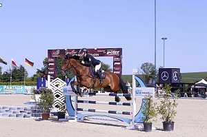 Ein Reiter springt mit einem braunen Pferd über ein blau-weißes Hindernis auf dem Sandplatz beim Reitturnier in Immenrode. Hinter dem Sprung sind der Begrüßungsbogen, Sponsorenbanner und wehende Länderflaggen zu sehen.