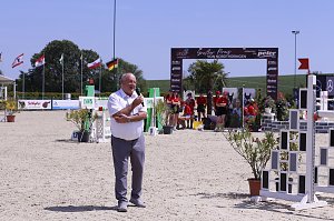 Helmut Peter spricht mit einem Mikrofon auf dem Springplatz beim Reitturnier in Immenrode. Hinter ihm stehen Helfer in roten Shirts vor dem Begrüßungsbogen und wehenden Fahnen.