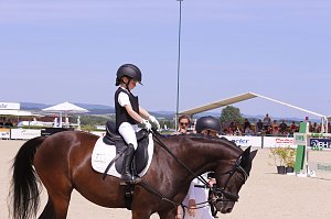 Ein Kind in weißer Reitkleidung und schwarzer Weste sitzt auf einem braunen Pferd auf dem Turnierplatz in Immenrode. Vor dem Pferd führen zwei Erwachsene das Tier, im Hintergrund sind Zuschauertribünen und Hindernisse zu sehen.
