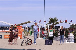 Siegerehrung beim Reitturnier in Immenrode: Auf dem Sandplatz stehen fünf Erwachsene, einer spricht ins Mikrofon. Vor ihnen liegen Preise wie ein Kinderfahrrad und Geschenktaschen, im Hintergrund sitzen Zuschauer auf den Tribünen unter wehenden Fahnen.