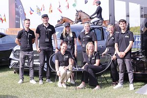 Gruppenfoto des Teams vom Autohaus Peter beim Reitturnier in Immenrode: Acht Teammitglieder in schwarzen Polohemden posieren vor ausgestellten Mercedes-Benz Fahrzeugen, im Hintergrund reiten Teilnehmer auf braunen Pferden über den Turnierplatz.
