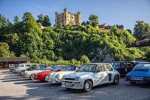 Reihe von klassischen Autos, darunter ein weißer Renault 5 Turbo mit Startnummer 52, ein roter und ein cremefarbener Porsche, die auf einem Parkplatz vor einer grünen bewaldeten Anhöhe stehen. Im Hintergrund thront das gelbe Schloss Hohenschwangau in Bayern unter blauem Himmel. Die Szene wirkt sonnig und freundlich.