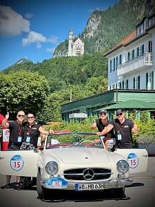Das Team vom Autohaus Peter steht vor einem weißen Mercedes-Benz Oldtimer mit Startnummer 15. Zwei Personen stehen links neben dem Auto, zwei rechts. Alle tragen schwarze Shirts, Sonnenbrillen und zeigen Daumen nach oben. Im Hintergrund ist Schloss Neuschwanstein auf einem bewaldeten Berg zu sehen, daneben ein weißes Gebäude mit grünen Fensterläden. Sonniger, klarer Tag.