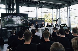 Podium mit sieben Mitgliedern der Geschäftsführung und Ausbildungsleitung der Autohaus-Peter-Gruppe bei der Begrüßung der neuen Auszubildenden. Davor sitzen Azubis in schwarzen T-Shirts und hören zu.