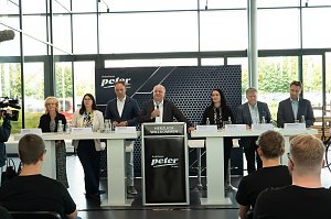 Podium mit sieben Mitgliedern der Geschäftsführung und Ausbildungsleitung der Autohaus-Peter-Gruppe bei der Begrüßung der neuen Auszubildenden. Davor sitzen Azubis in schwarzen T-Shirts und hören zu.
